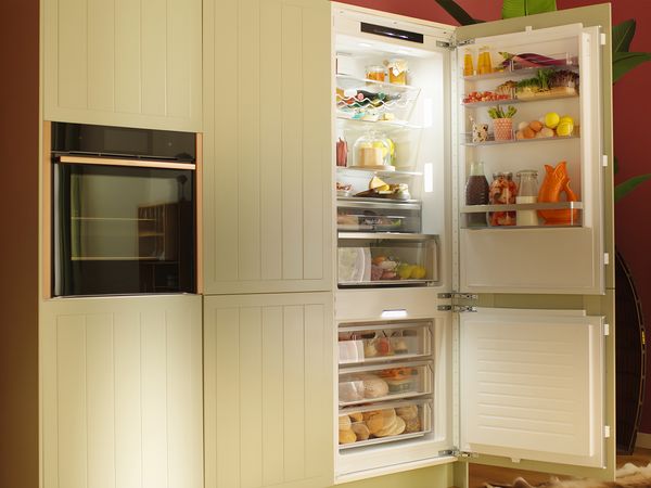 Un frigorifero elegante di colore verde chiaro con la porta aperta e una serie di alimenti e bevande disposti in modo ordinato al suo interno, con una parete di colore rosso sullo sfondo.