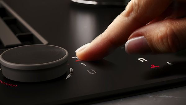 Primo piano di una mano che preme un pulsante sui comandi di un piano cottura moderno, con una manopola rotonda e una serie di indicatori digitali.