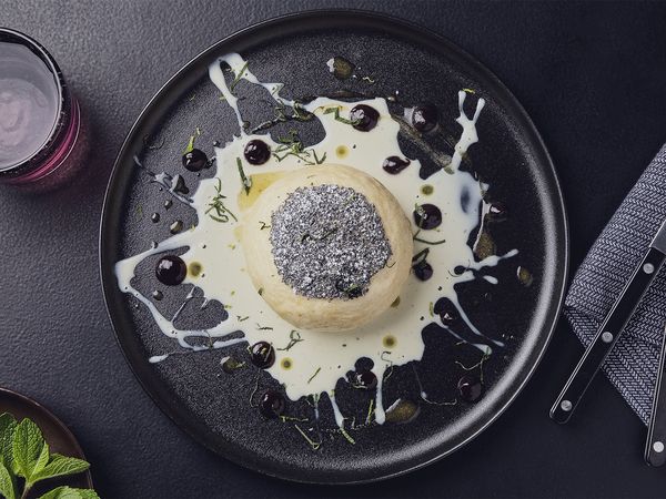 Single yeast dumpling topped with poppy seeds and powdered sugar, surrounded by vanilla sauce and plum syrup on a black plate.