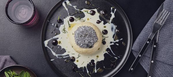 Single yeast dumpling topped with poppy seeds and powdered sugar, surrounded by vanilla sauce and plum syrup on a black plate.