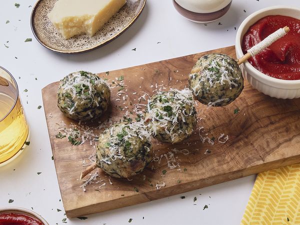 Four spinach and cheese dumplings on a wooden board topped with grated cheese and parsley, served with a bowl of tomato-date sauce and rustic bread on the side.