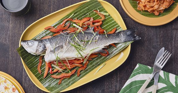 Ein ganzer gedämpfter Wolfsbarsch, angerichtet auf einem Bananenblatt mit in Scheiben geschnittenen roten Paprikaschoten und Frühlingszwiebeln, präsentiert auf einem gelben Teller mit Kimchi als Beilage.