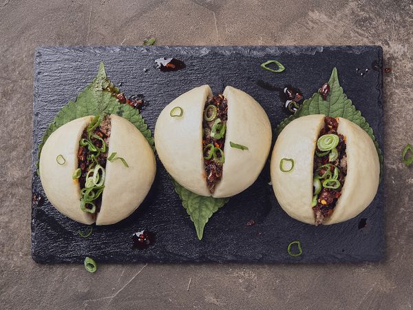 Three steamed pork buns sliced open and filled with spiced ground meat, topped with sliced scallions and chili crisp, served on shiso leaves over a black slate plate.