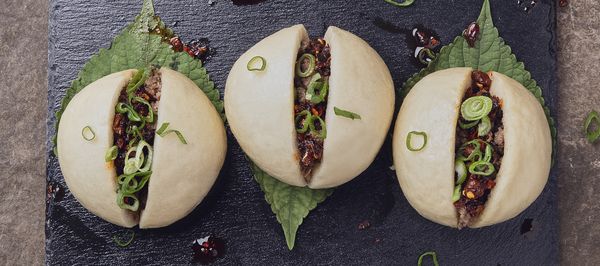 Three steamed pork buns sliced open and filled with spiced ground meat, topped with sliced scallions and chili crisp, served on shiso leaves over a black slate plate.