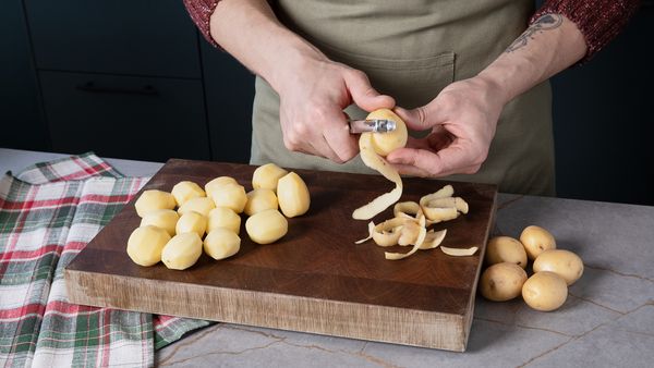 Person peels potatoes at a kitchen counter, with peeled potatoes on a wooden chopping board and a festive tablecloth to the side.