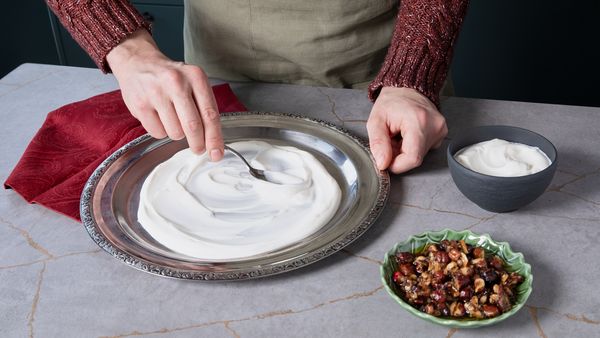 Yogurt being spread onto a silver serving plate, with a bowl of hazelnuts and a red napkin beside it.