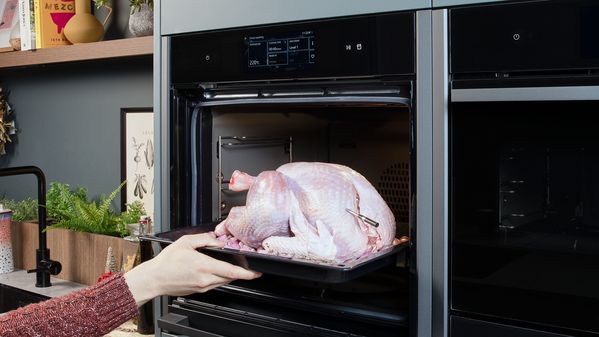 A whole raw turkey with a food thermometer on a baking tray being slid into an open oven.
