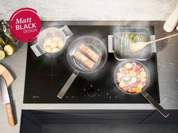 NEFF Induktionskochfeld im Matt Black Design mit Twist Pad® Bedienung, belegt mit Töpfen und Pfannen voller Gemüse, Fisch und Kartoffeln.