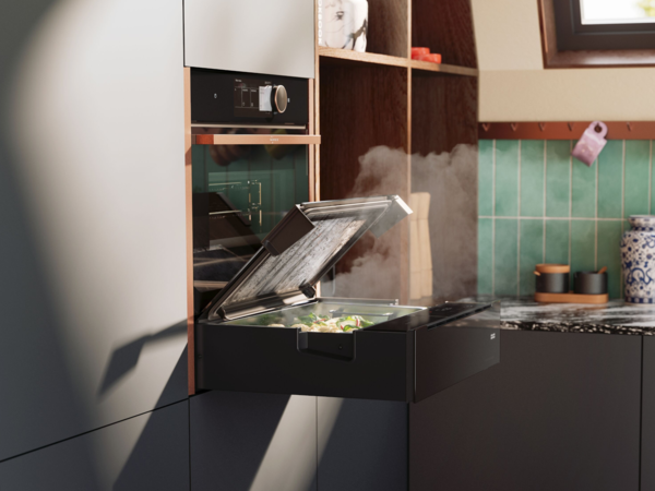 NEFF Dampfschublade in moderner Küche – platzsparend unter einem Backofen eingebaut, mit geöffnetem Glasdeckel und aufsteigendem Dampf beim Garen von frischem Gemüse.