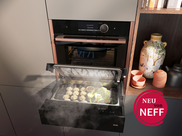 NEFF Dampfschublade in moderner Küche – platzsparend unter einem Backofen eingebaut, mit geöffnetem Glasdeckel und aufsteigendem Dampf beim Garen von frischem Gemüse.
