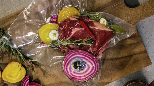 Вакуумно запечатан пакет със сурово говеждо месо, ярки зеленчуци, билки и ядливи цветя върху дървена дъска.