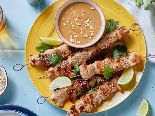 Chicken satay skewers with peanut sauce, garnished with lime and cilantro, on a yellow plate.