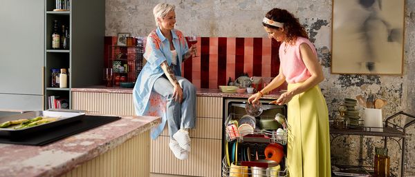 Due donne in cucina, una &egrave; seduta sul piano di lavoro con un bicchiere in mano, mentre l&rsquo;altra sta svuotando la lavastoviglie