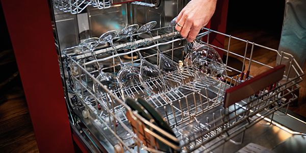 Käsi asetab veiniklaasi nõudepesumasina ülemisse korvi, mis on täis klaase ja nõusid.