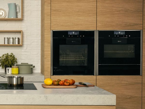 Модерна кухня с две елегантни черни фурни и дървена дъска за рязане с пресни домати и лимон.