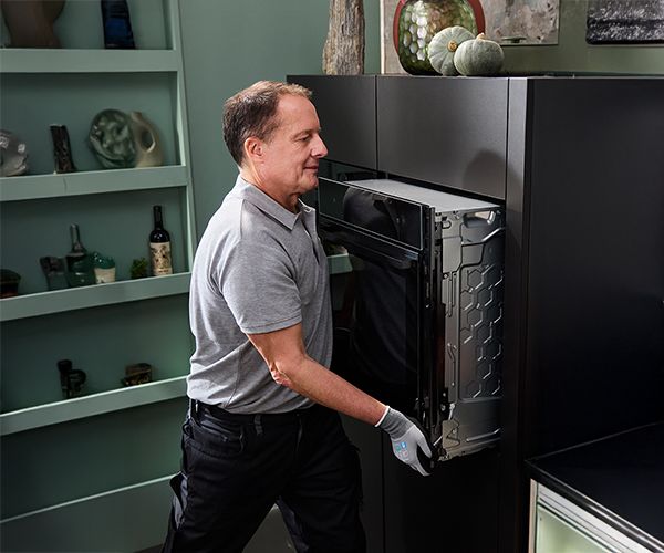 
       Un technicien installe un appareil de cuisine encastrable, portant des gants et une chemise grise, dans une cuisine moderne d&eacute;cor&eacute;e avec soin.
      