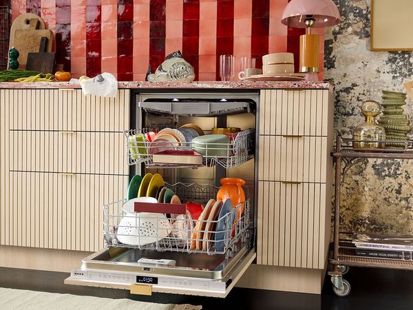 Стилна кухня с частично отворена съдомиялна машина, пълна с цветни чинии, разположена на фона на яркочервен гръб с плочки.