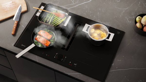 A sleek black induction cooktop is surrounded by fresh vegetables, a pot of lemons, and kitchen tools, set in a modern kitchen environment.