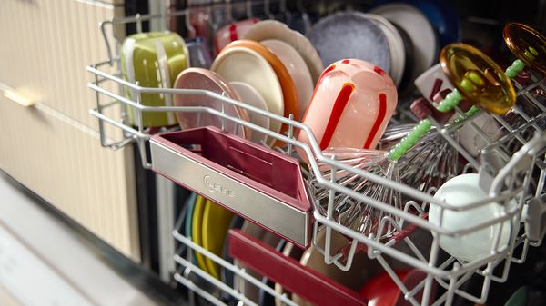 Частично заредена съдомиялна машина показва разнообразие от цветни чинии, чаши и прибори, подредени по рафтовете си.