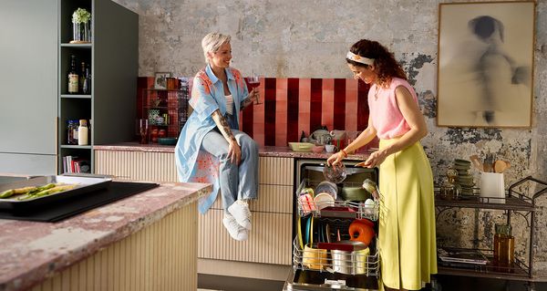 Twee vrouwen in een stijlvolle keuken: de ene zit op het aanrecht met een drankje, de andere laadt de vaatwasser.