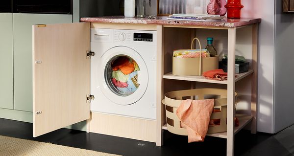 Зона за пране с пералня зад отворена дървена врата на шкаф, цветно пране вътре и организирано място за съхранение отдолу