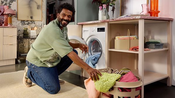 Човек коленичи до кош за пране, сортирайки цветни дрехи близо до пералня в стилно перално помещение.