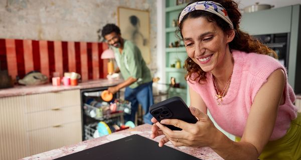 En kvinna i en rosa tröja använder en smartphone medan en person i bakgrunden plockar ur en diskmaskin i ett stilfullt kök.