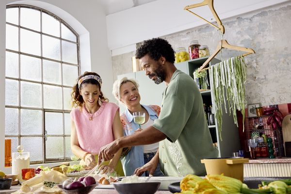 Tre amici sorridenti mentre preparano il cibo insieme in una cucina luminosa e accogliente con ingredienti freschi sul bancone.