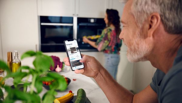 People in kitchen one putting food in oven and one using the Home Connect app on mobile phone