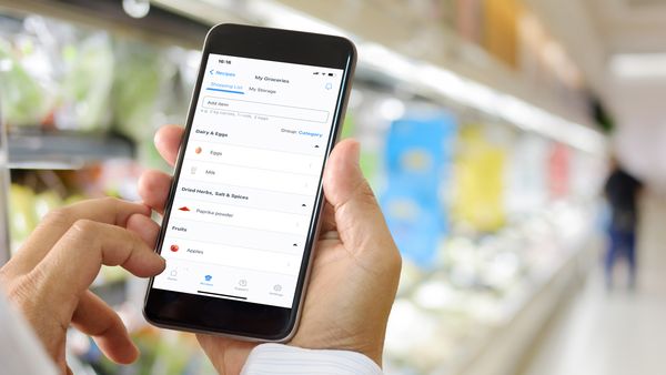 Zwei H&auml;nde halten ein Smartphone hoch, auf dem die Home Connect-App zu sehen ist. Im Hintergrund ist der K&uuml;hlregalbereich eines Supermarkts zu sehen.