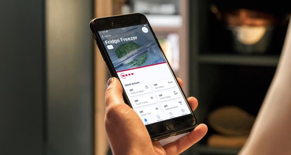 Eine Nahaufnahme einer Hand, die ein Smartphone h&auml;lt, auf dem die Home Connect-App zu sehen ist, sowie ein K&uuml;hlschrank im Hintergrund.