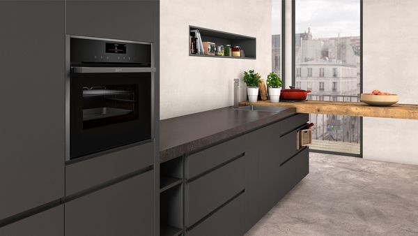 Cucina moderna con mobili scuri, forno incorporato, piano di lavoro in legno, erbe aromatiche in vaso e vista sugli edifici cittadini attraverso ampie finestre.