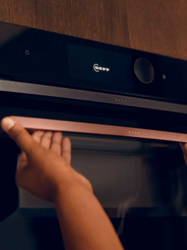 K&auml;det n&auml;ytt&auml;v&auml;t harjatun pronssisen kahvan antrasiitinharmaan NEFF-uunin edess&auml;.