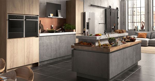 Cucina open space con isola centrale, finiture in cemento e legno, elettrodomestici integrati e piano di lavoro con cibi