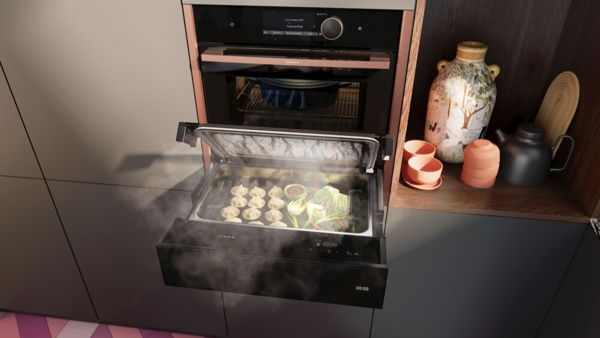 Een moderne keukensc&egrave;ne met een stoomlade gevuld met dumplings en groenten, naast decoratieve keramiek en boeken.