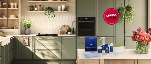 Cuisine moderne et lumineuse avec suspensions jaunes, armoires vertes, plantes en pot et plan de travail gris avec ustensiles. 