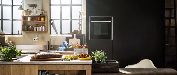 Кухня с високи сиви шкафове с вградена фурна Neff. Кухненски остров с печено месо върху дъска и различни пресни зеленчуци върху плота.