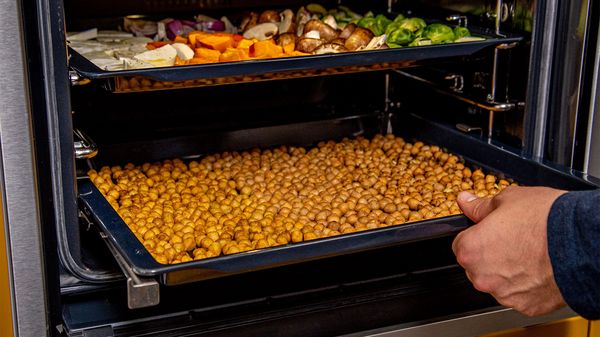 Forno aperto con teglia di verdure tagliate sul ripiano superiore e mano che inserisce un&rsquo;altra teglia con ceci