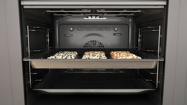 A close-up view inside an oven with a tray of three rectangular food items, each topped with different seeds.