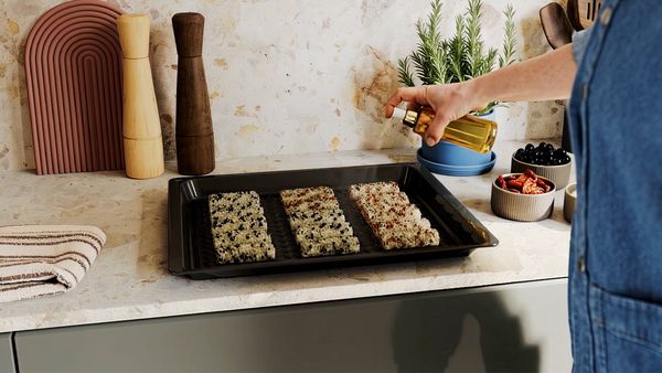 A person's hand is spraying oil onto a tray of three rectangular food items topped with seeds, on a kitchen counter.