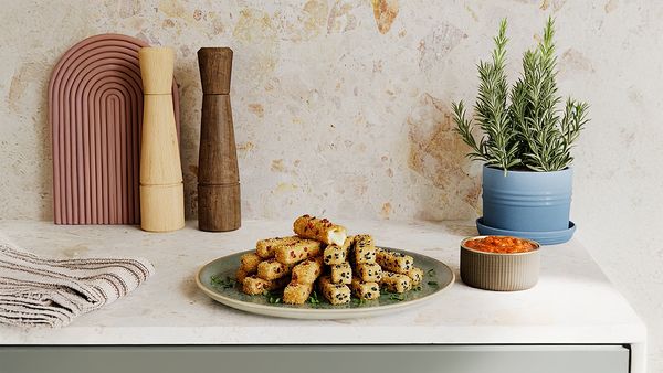  A plate of golden-brown, breaded cheese sticks, some topped with seeds, sits on a kitchen counter next to a bowl of sauce and a potted plant.