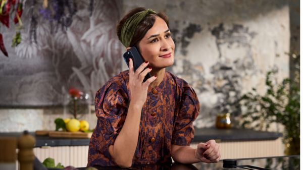 Eine Person steht in einer modernen K&uuml;che, telefoniert und kocht, umgeben von Kochutensilien und Zutaten.