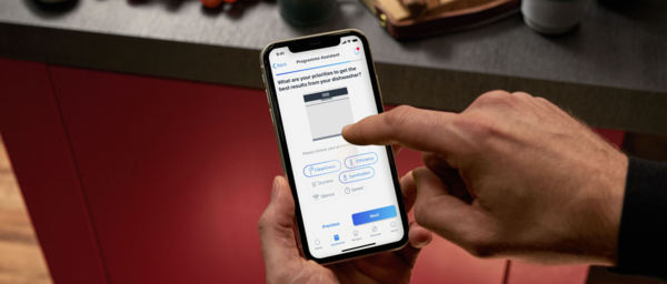 Eine Frau steht in einer Designerk&uuml;che mit offenem Geschirrsp&uuml;ler und anderen Einbauger&auml;ten und blickt auf ihr Smartphone