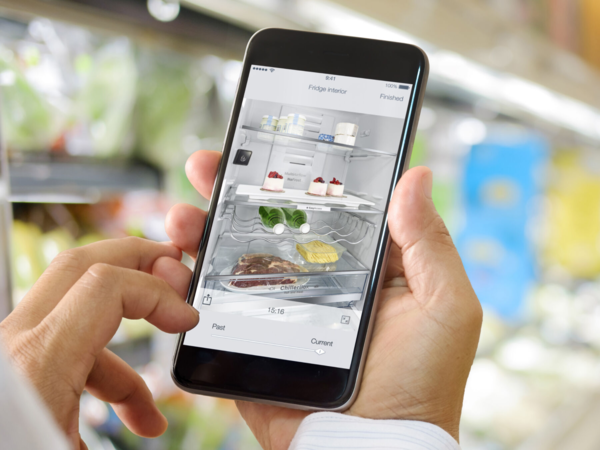 Eine Person h&auml;lt ein Smartphone mit einer App f&uuml;r den K&uuml;hlschrank, vor dem Hintergrund einer modernen K&uuml;che und einem offenen K&uuml;hlschrank voller Lebensmittel.