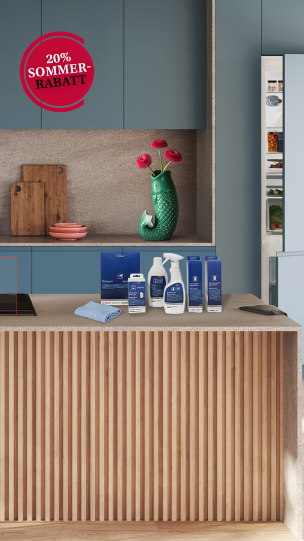 Moderne K&uuml;che mit blauen Schr&auml;nken, einer K&uuml;cheninsel aus Holz und Rattanhockern. Auf der Arbeitsfl&auml;che befinden sich frische Fr&uuml;chte, Getr&auml;nke und Reinigungsmittel. Das Bild zeigt au&szlig;erdem einen Sommerrabatt von 20 %.