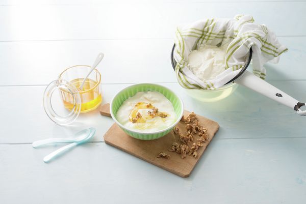 Ciotola di yogurt con miele e noci su tagliere, miele in brocca, yogurt filtrato in colino e cucchiaini sul tavolo