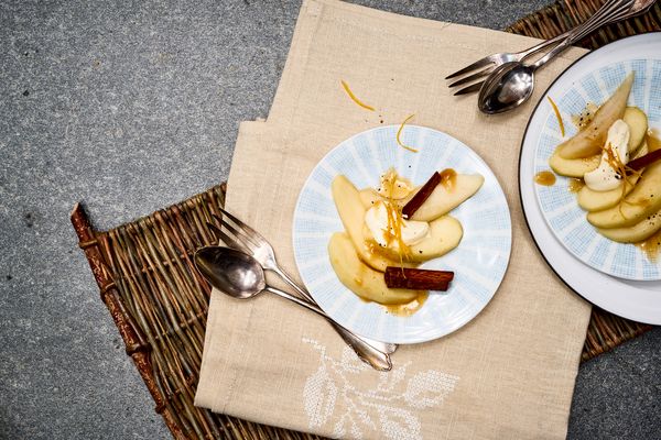 Piatti di pere cotte conditi con panna, bastoncini di cannella e scorza d'arancia, disposti su un tappetino intrecciato e un panno beige.