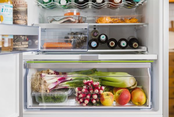 Interno del frigorifero con ripiani e cassetto riempiti con bottiglie, vaschette, frutta e verdura fresca