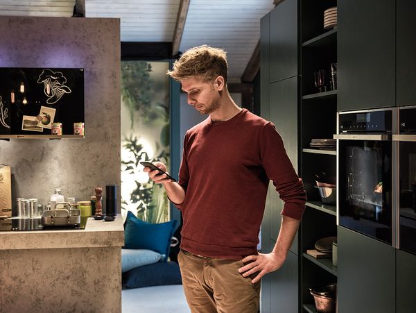Man using Home Connect app on mobile phone in kitchen with NEFF built-in appliances