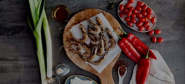 Εικόνα φρέσκων γαρίδων σε ένα ξύλο κοπής, περιτριγυρισμένων από ντοματίνια, κόκκινες πιπεριές, πράσινα κρεμμύδια, ελαιόλαδο και μπαχαρικά.